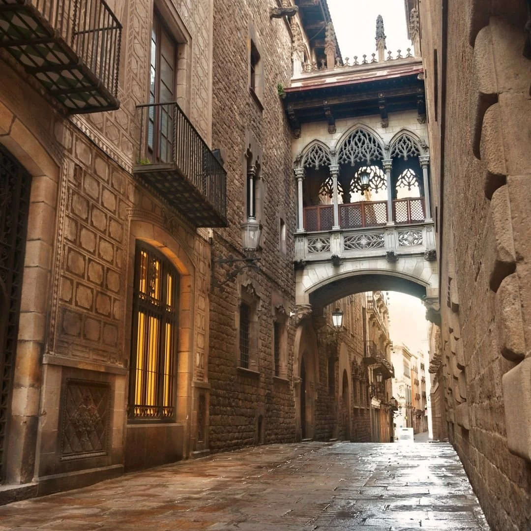A quiet historic street in Spain with stone buildings and iron balconies, where a Gothic-style bridge connects two façades above a narrow, empty lane washed in soft daylight.