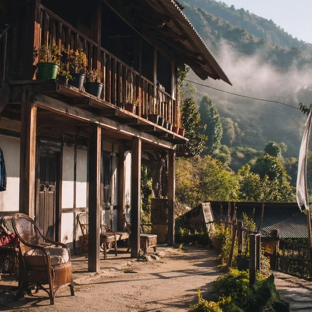 Traditional wooden homestay on a mountain slope in Arunachal Pradesh, with warm sunlight, mist drifting through lush greenery, and simple lived-in outdoor seating creating a calm, grounded atmosphere