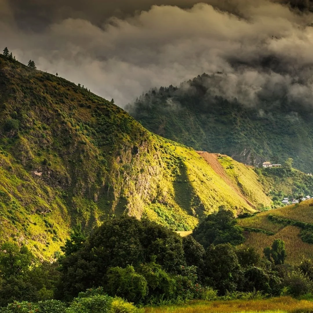 Sunlit green hills in Arunachal Pradesh with misty clouds drifting over layered mountains and a small hillside village in the distance