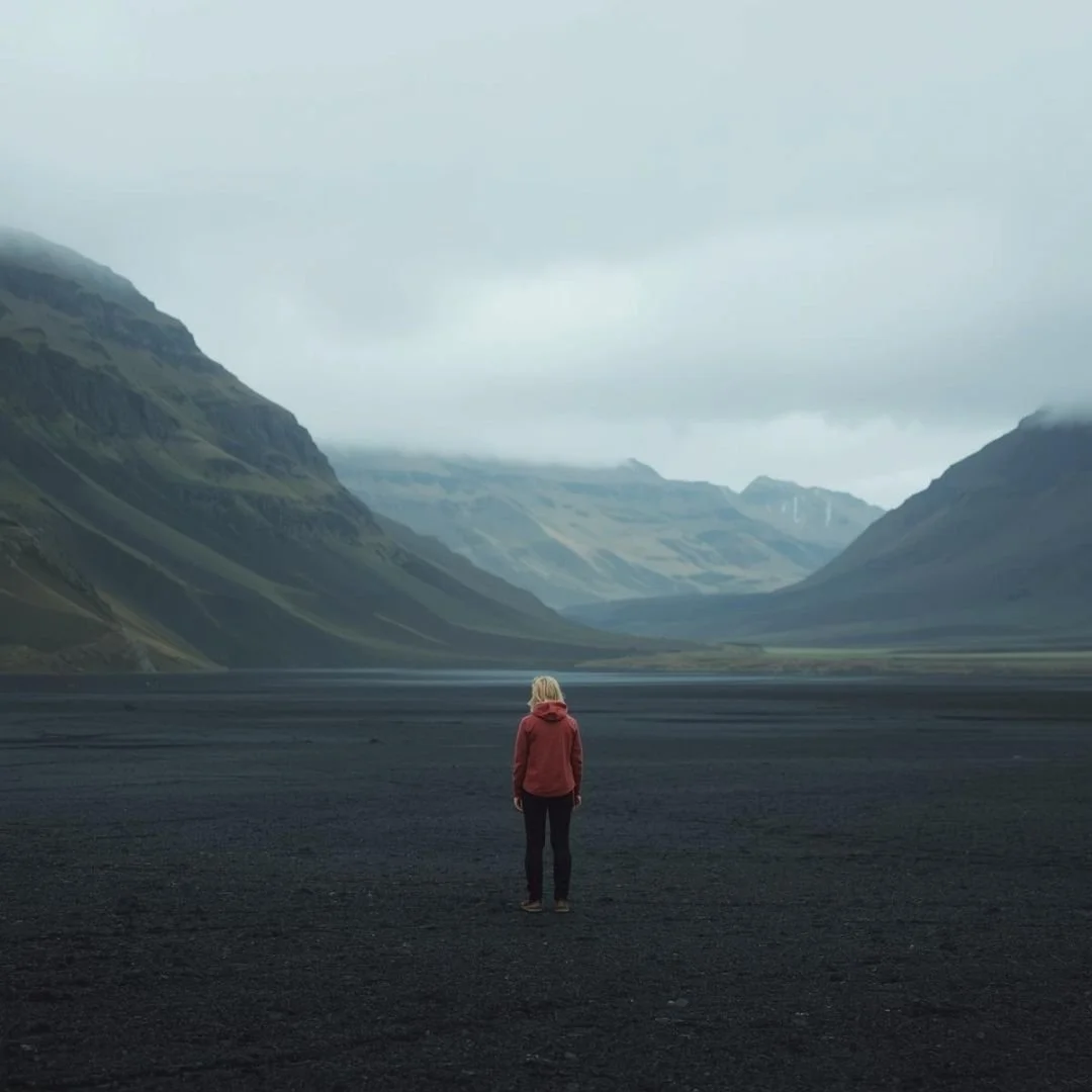 A lone traveller standing on a vast black sand plain in Iceland, facing a wide valley framed by green mountains beneath a heavy, overcast sky, capturing a feeling of solitude and scale.