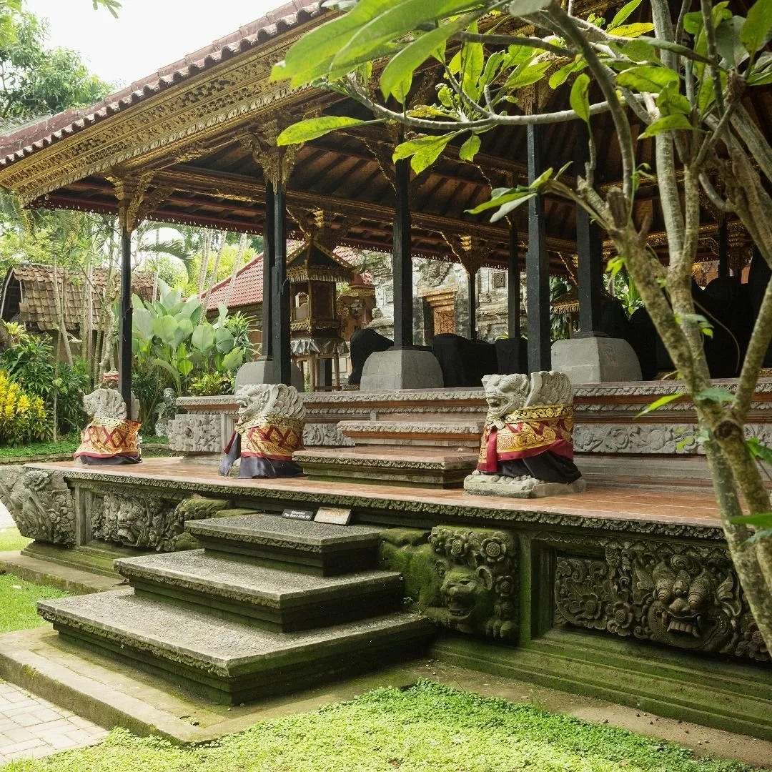 A traditional Balinese temple pavilion with carved stone steps, guardian statues draped in ceremonial cloth, and lush garden surroundings, reflecting Bali’s living spiritual architecture.