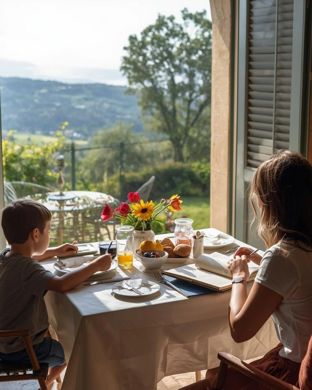 Parent and child sharing a relaxed breakfast at a sunlit table by an open window, with fresh flowers, fruit, and a countryside view outsid