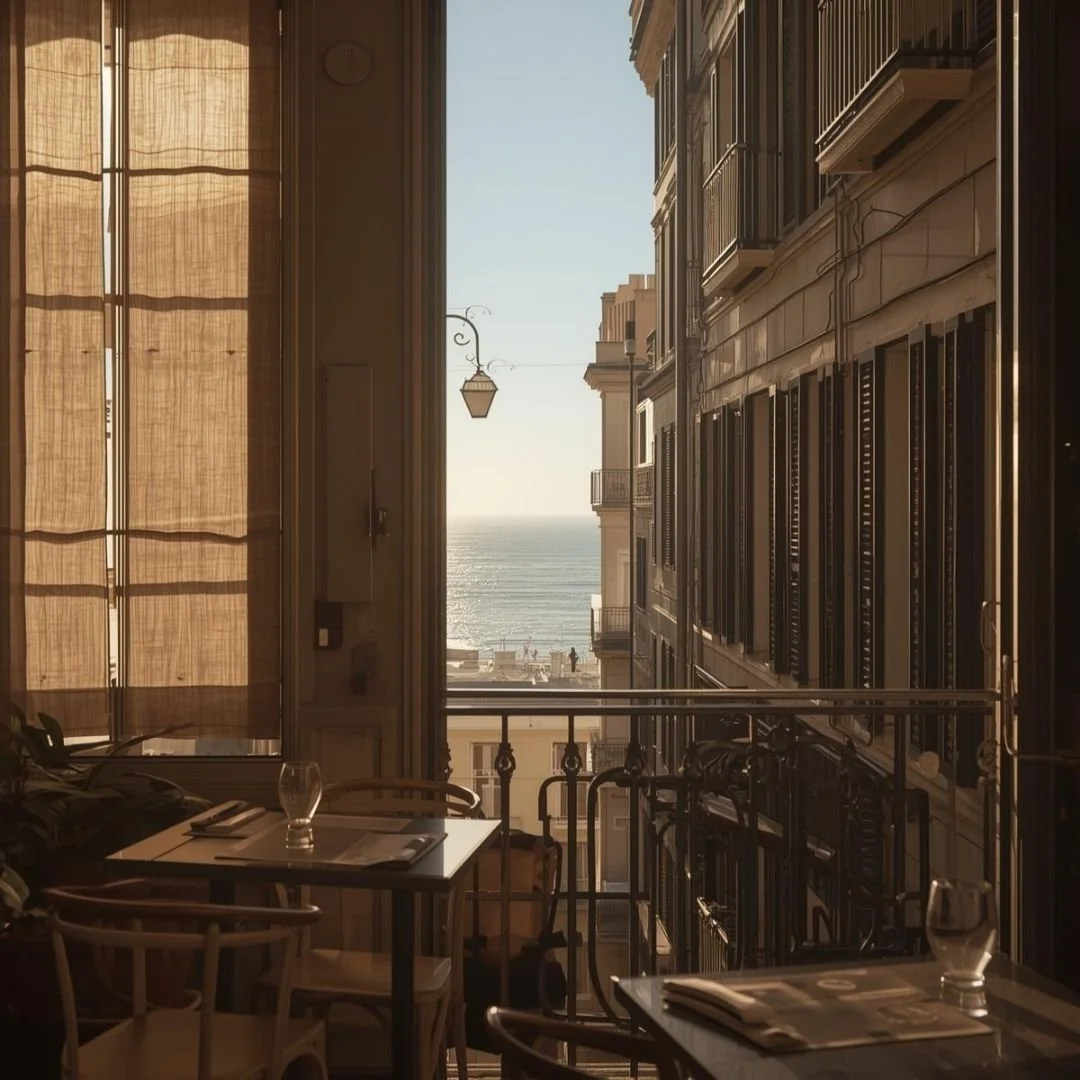 Soft morning light filters through linen curtains onto a small café table, opening to a narrow Barcelona balcony with shuttered buildings on either side and a quiet glimpse of the sea beyond, calm and unhurried.