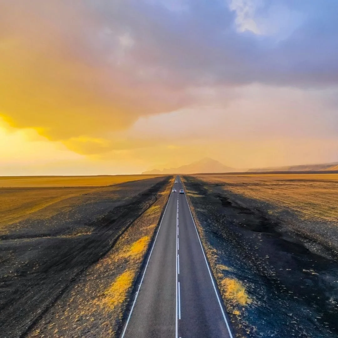 A long straight road cutting through Iceland’s open plains at sunset, with golden light washing over volcanic terrain and distant mountains fading into the horizon.