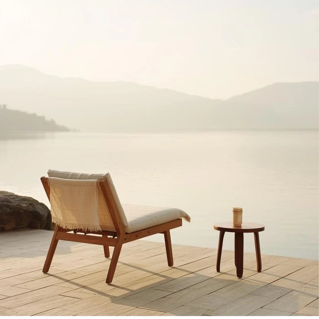 Wooden lounge chair and small table with a takeaway coffee on a quiet lakeside terrace, with misty mountains in soft morning light.