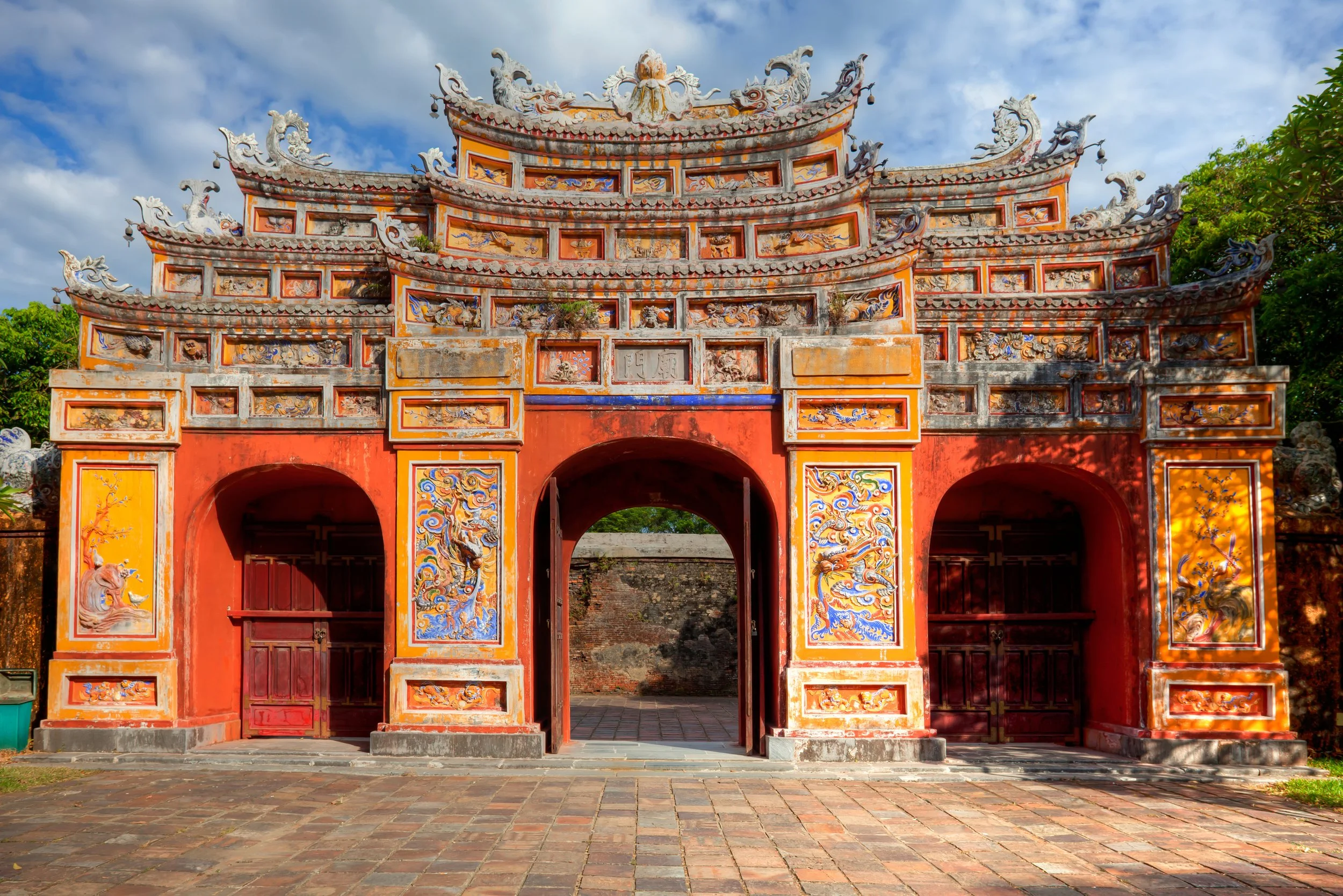 Colorful historic Chinese gate with ornate sculptures and painted details, set against a partly cloudy sky with trees in the background.