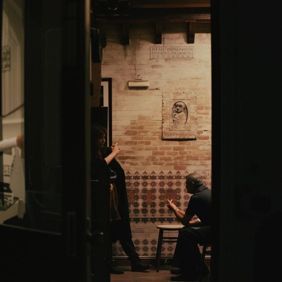 Intimate Czech interior with brick walls and vintage artwork, two people in quiet conversation, low light and warm tones creating a reflective, lived-in atmosphere.