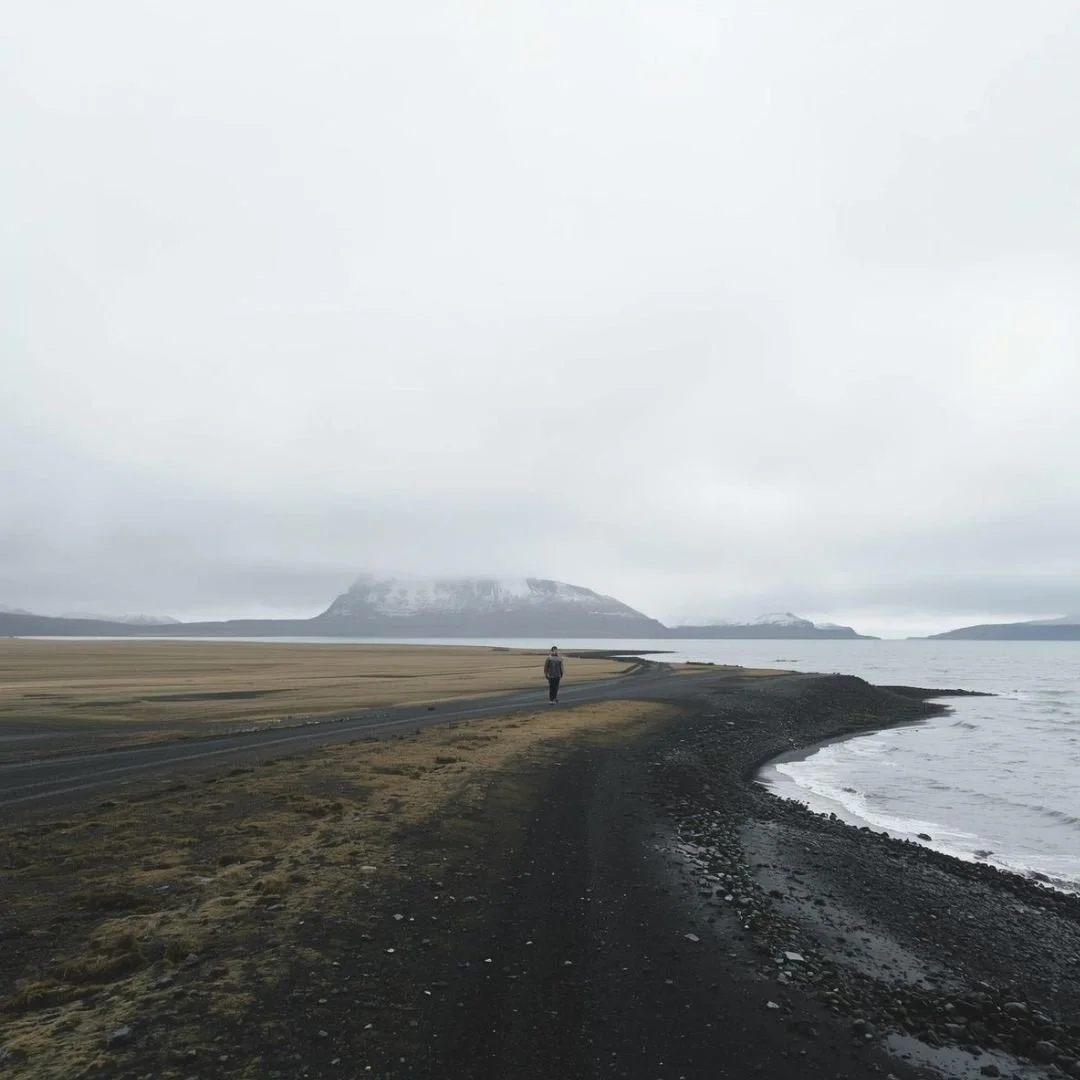A solitary traveller walking slowly along a narrow coastal road in Iceland, with black sand, still water, and distant mountains under a pale overcast sky, the landscape vast and quiet around them.