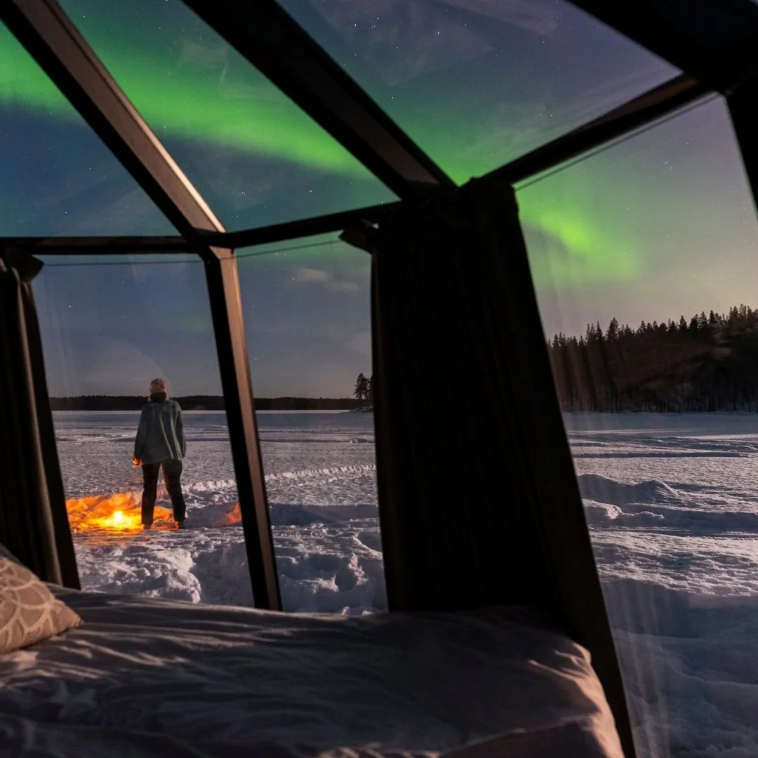 View from a glass-roof cabin overlooking a snowy landscape, with a person standing by a fire under the northern lights.