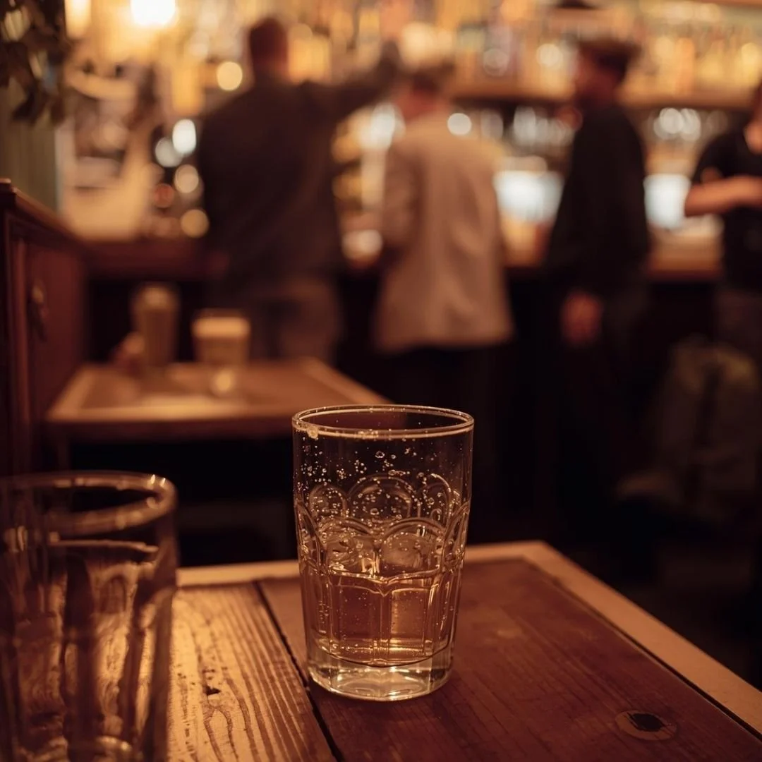 A half-filled glass resting on a wooden table inside a warmly lit European bar, with blurred figures and shelves behind, evoking an intimate evening ritual and local atmosphere.