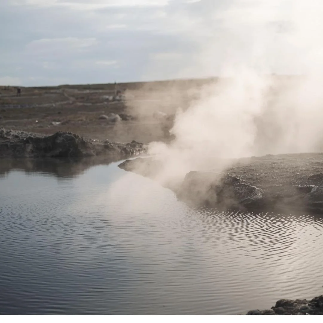 Steam rising gently from a geothermal pool in Iceland, where calm water meets dark volcanic earth under soft, muted daylight, creating a quiet, elemental atmosphere.