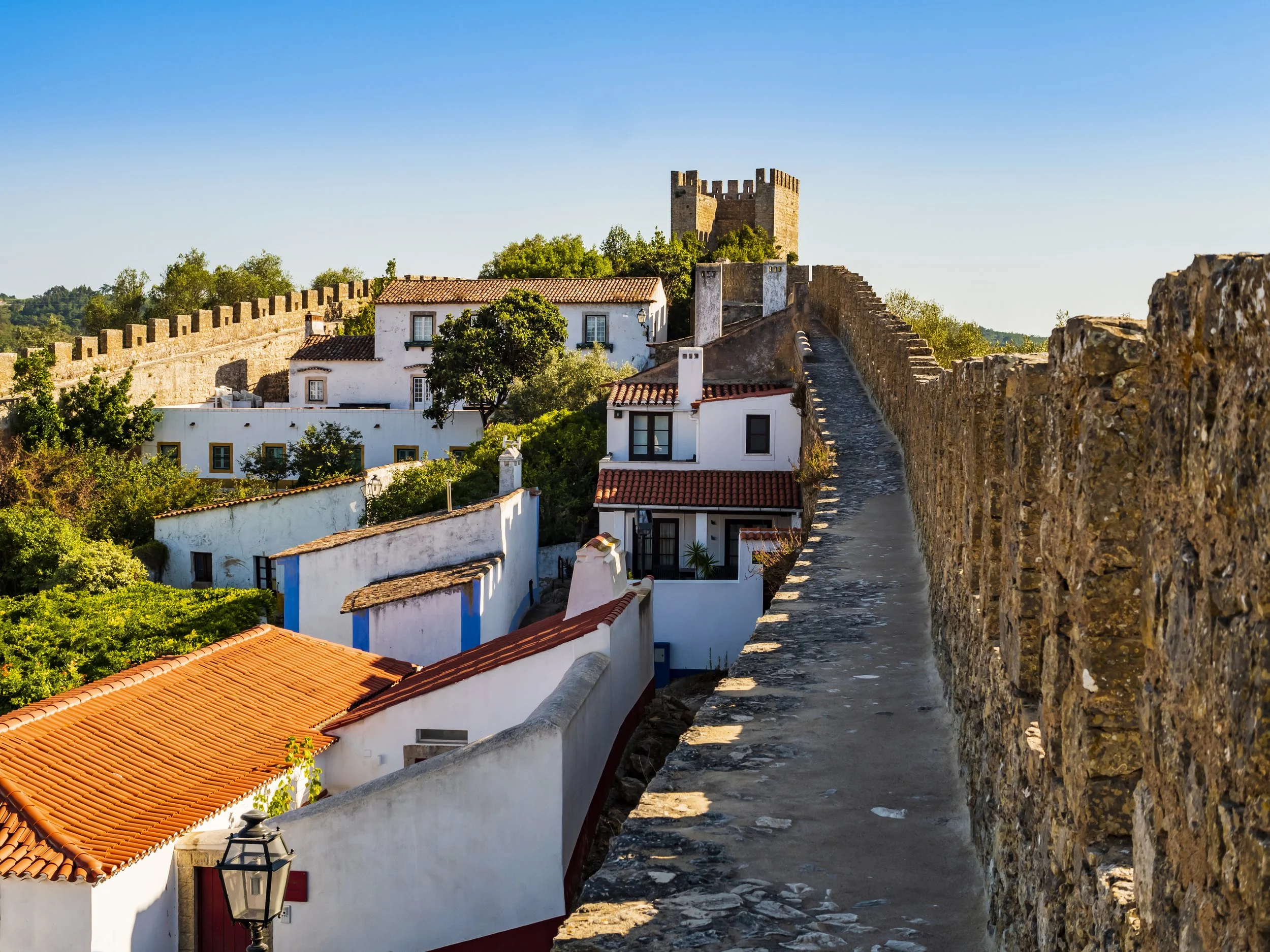 View of a historic stone wall with a walkway, overlooking white houses with red tile roofs and a castle in the background, under a clear blue sky.