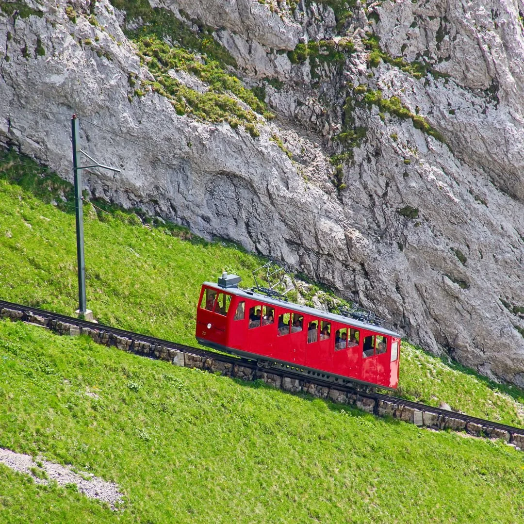 A red mountain cable car ascending a steep green grassy hillside with rocky terrain in the background.