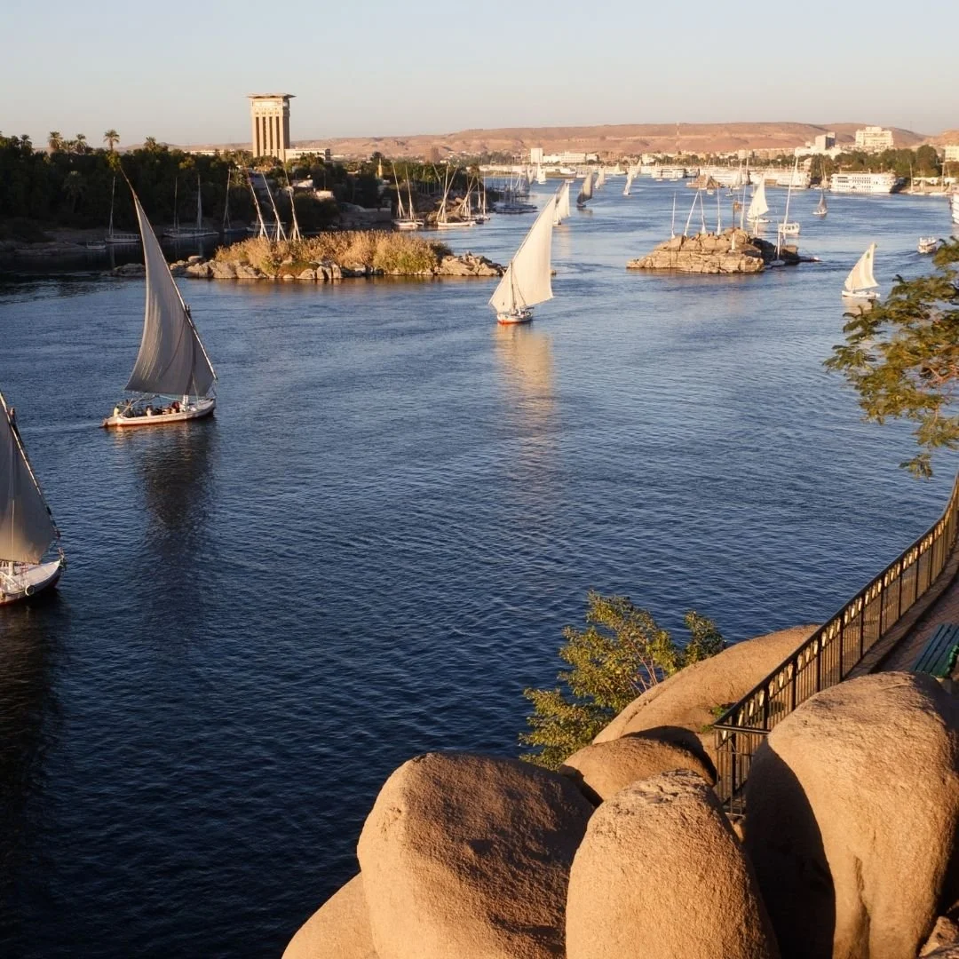 Felucca boats sailing on the Nile River in Egypt, with rocky riverbanks and distant shoreline visible.
