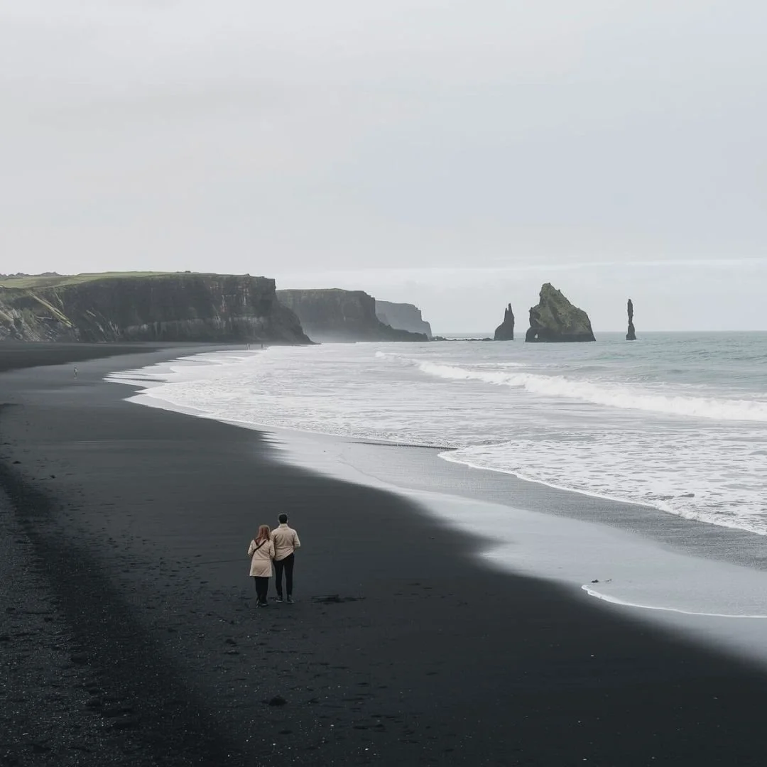A couple standing on Iceland’s black sand beach as gentle waves meet the shore, surrounded by cliffs and misty sea stacks.