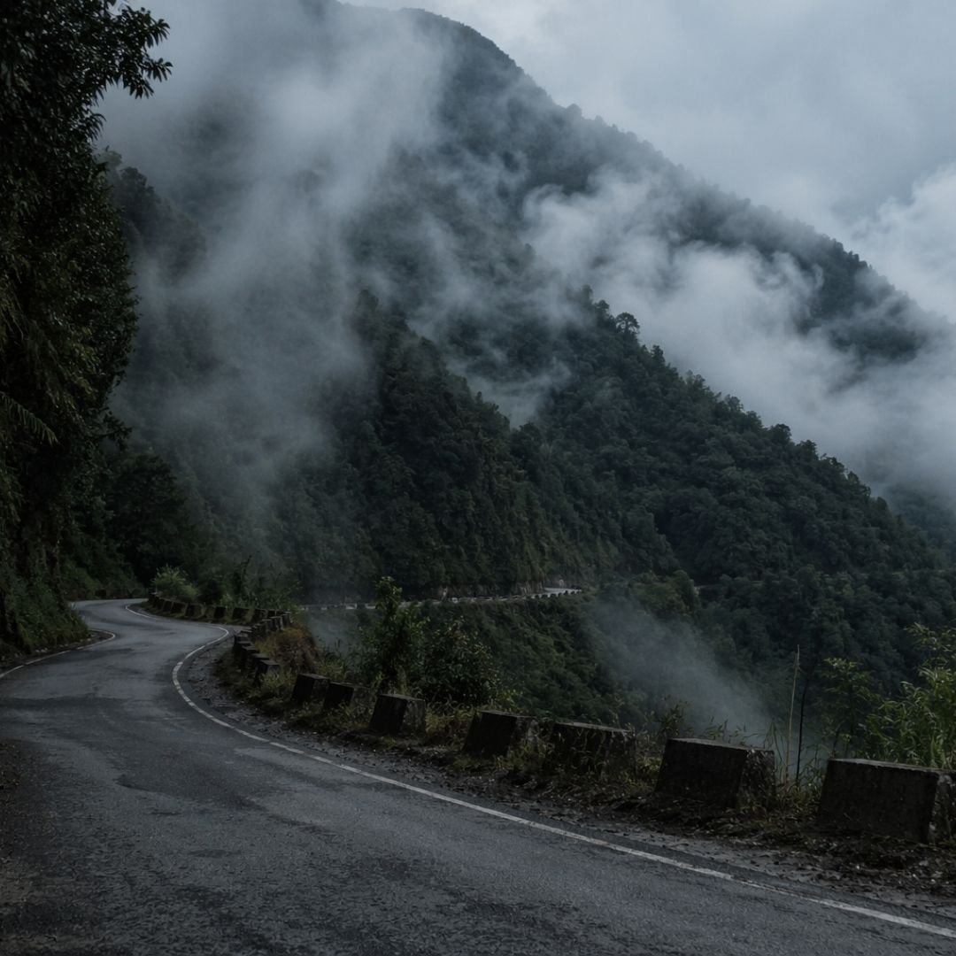 Winding mountain road in Arunachal Pradesh cutting through dense forest and mist, disappearing into low clouds, with damp textures and moody natural light creating a sense of isolation and quiet movement