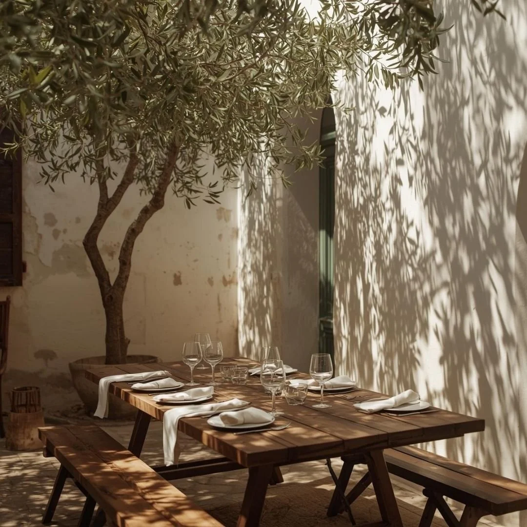 A shaded Andalusian courtyard with a wooden table set for a long lunch beneath an olive tree, sunlight casting soft leaf shadows on whitewashed walls.