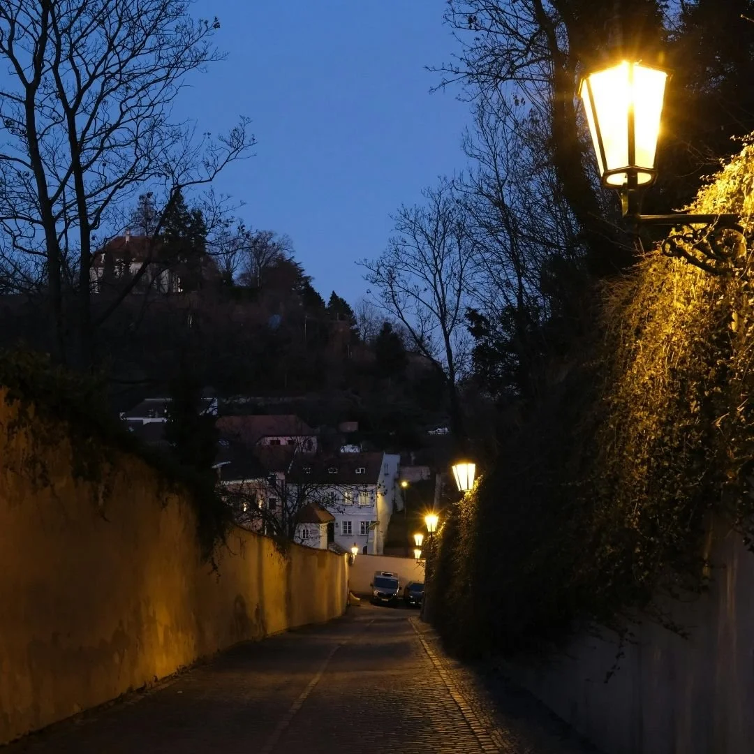 A quiet cobblestone lane at dusk, softly lit by warm street lamps, with bare trees and old town houses receding into the distance, creating an intimate, timeless atmosphere
