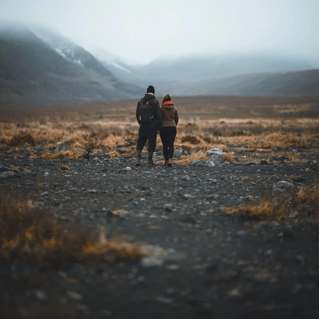 A couple walking away across a raw Icelandic landscape, surrounded by rocky ground and muted grass, with misty mountains in the distance under an overcast sky.