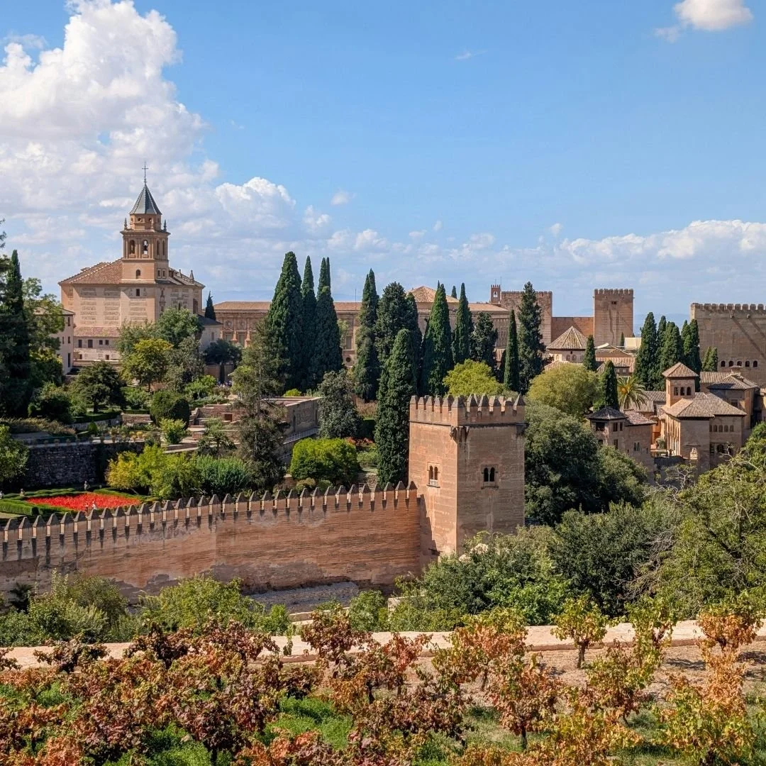 Sunlit view of the Alhambra in Granada, with terracotta fortress walls, towers, and cypress trees rising above lush gardens under a clear blue sky.