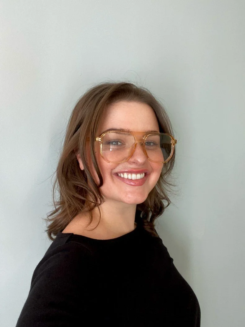 A young woman with shoulder-length brown hair and a tattoo on her left arm, smiling and looking over her shoulder outdoors with green trees and a wooden fence in the background.