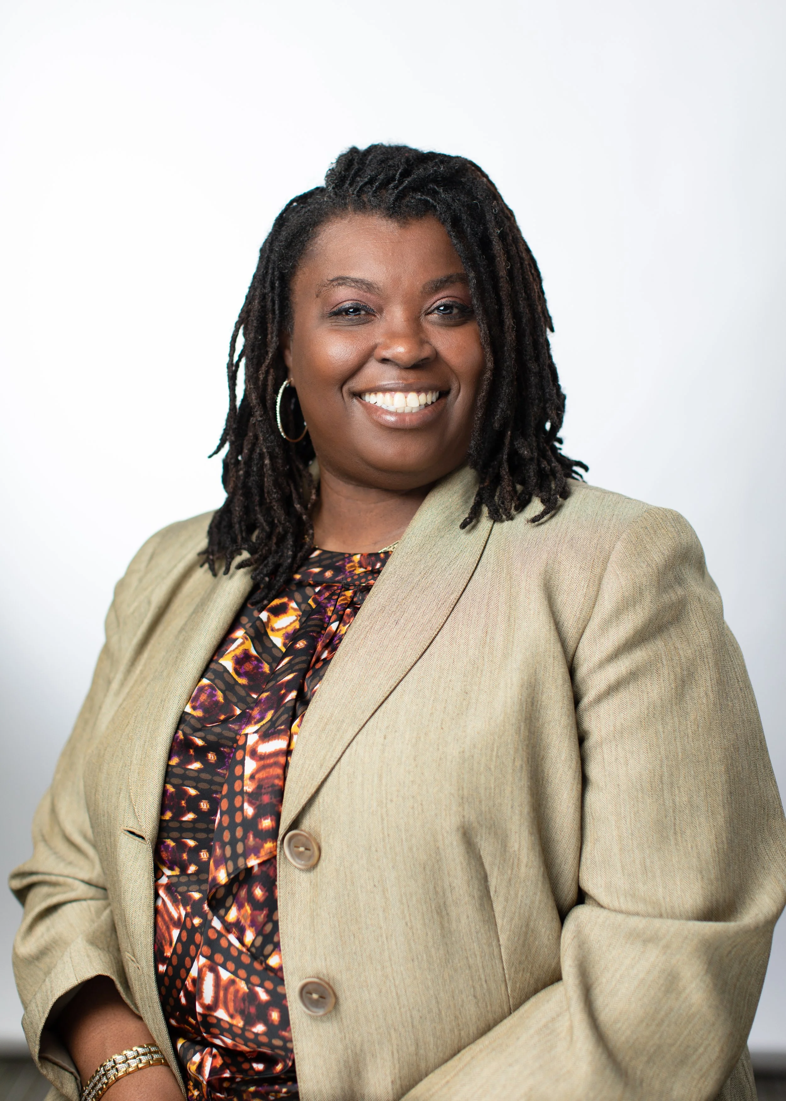 A woman with dark curly hair and hoop earrings smiling, dressed in a beige blazer over a colorful patterned blouse, standing against a plain white background.