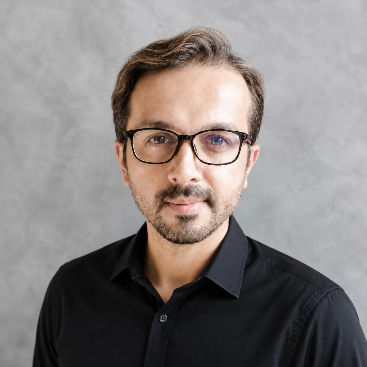 A man with brown hair, glasses, and a beard wearing a black shirt, standing against a gray background.