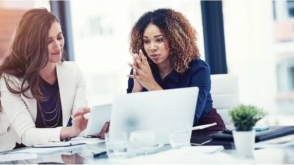Two women in a business meeting looking at a laptop and a tablet in a bright office.