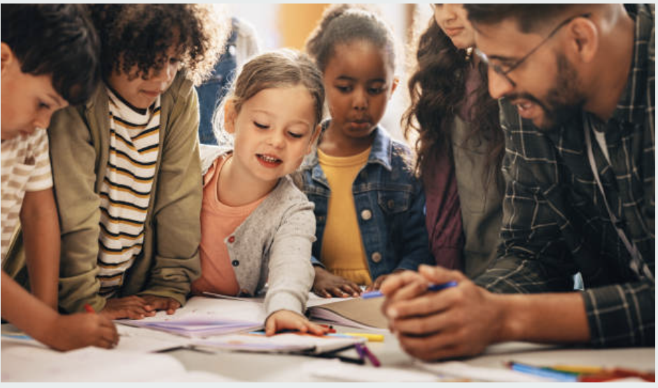 A group of children gathered around an adult, engaged in an activity or discussion, with papers and pens on the table.