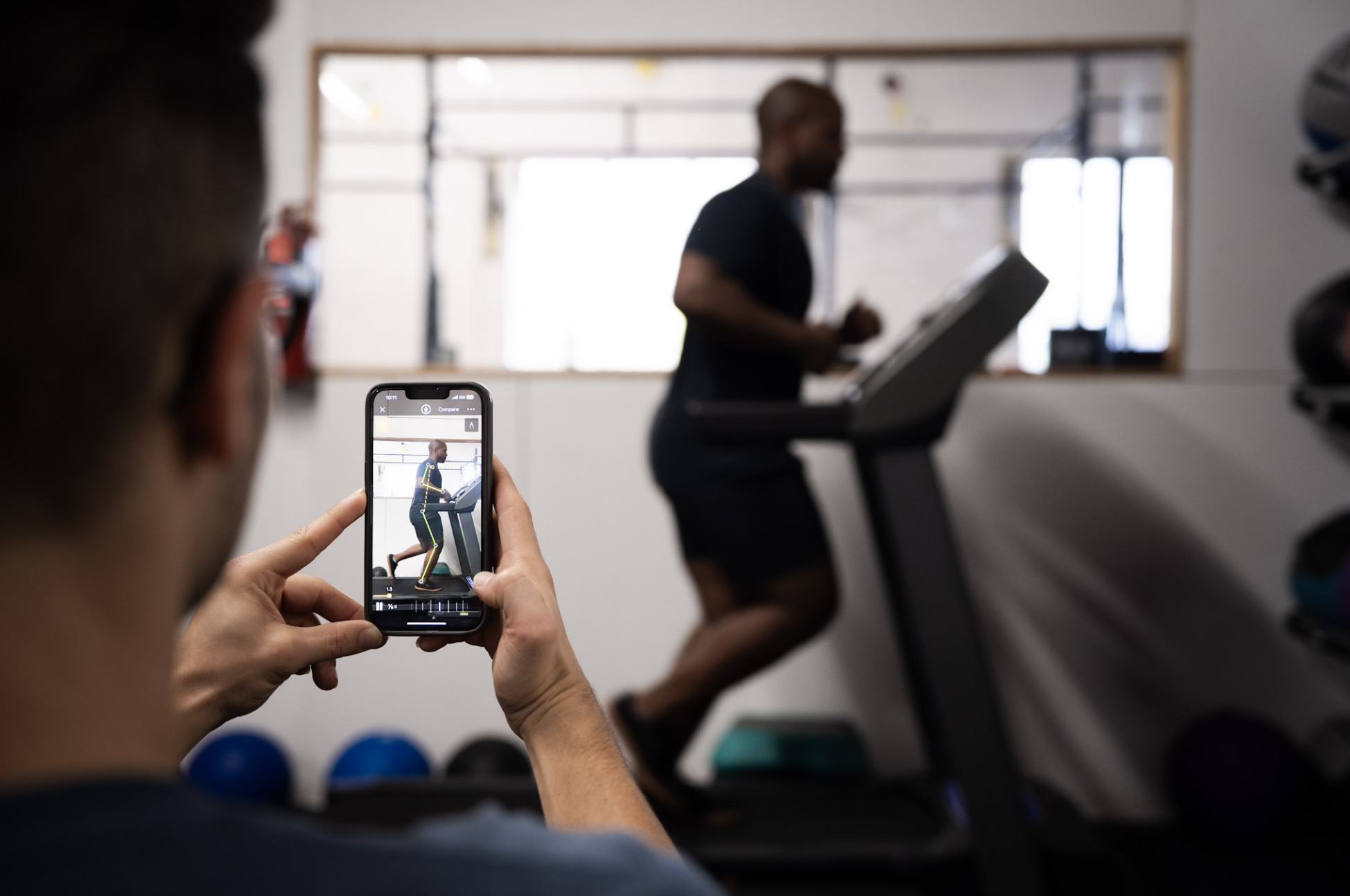 Person taking a photo of a man running on a treadmill with a smartphone in a gym.