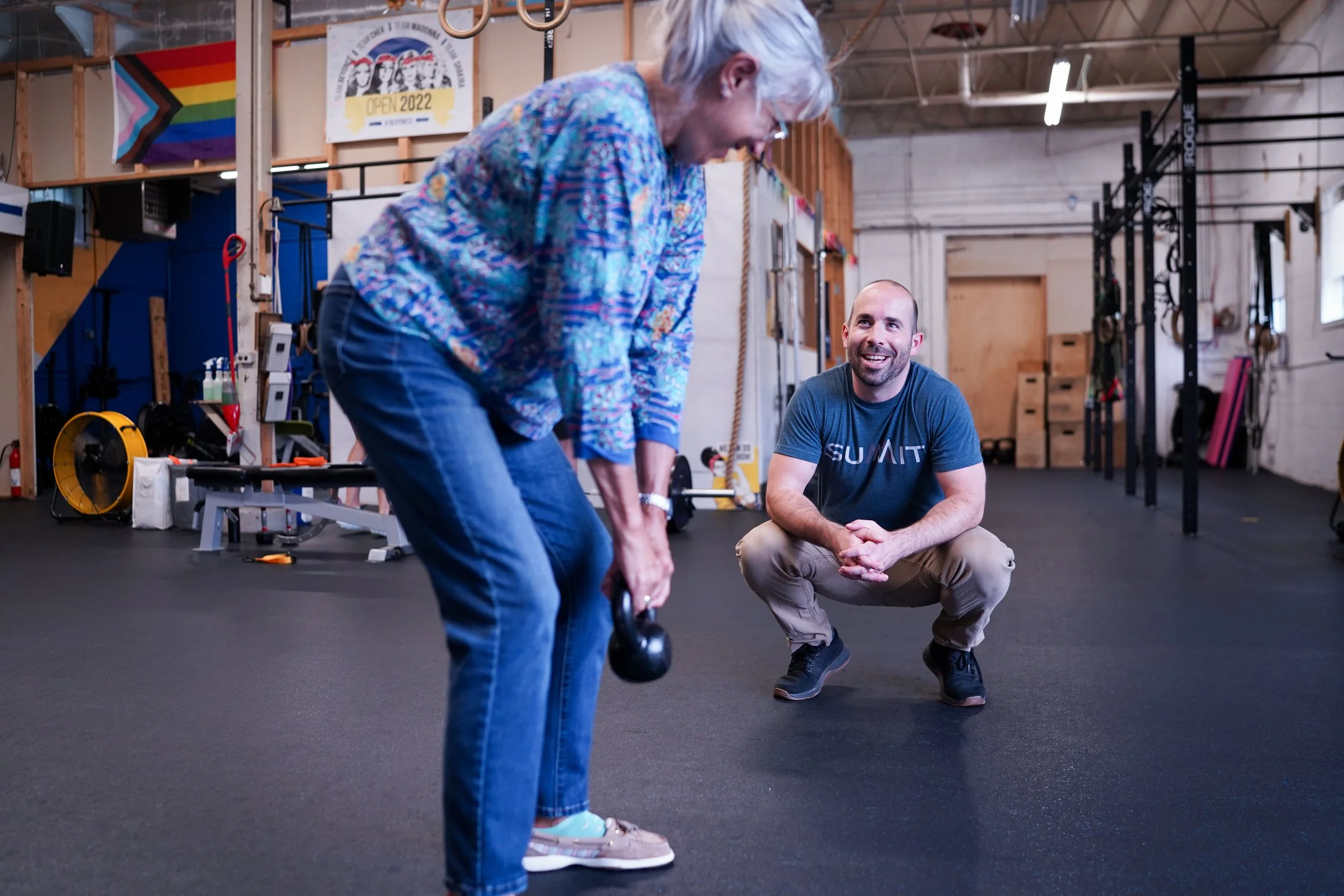 A woman lifting a kettlebell while a man crouches and observes in a gym.