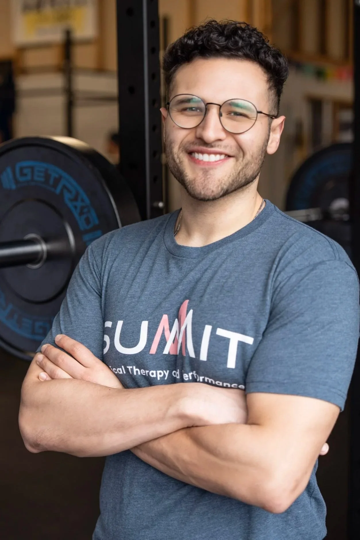 A smiling young man wearing glasses and a blue T-shirt with 'SUMMIT' printed on it, standing with arms crossed in a gym or training facility.