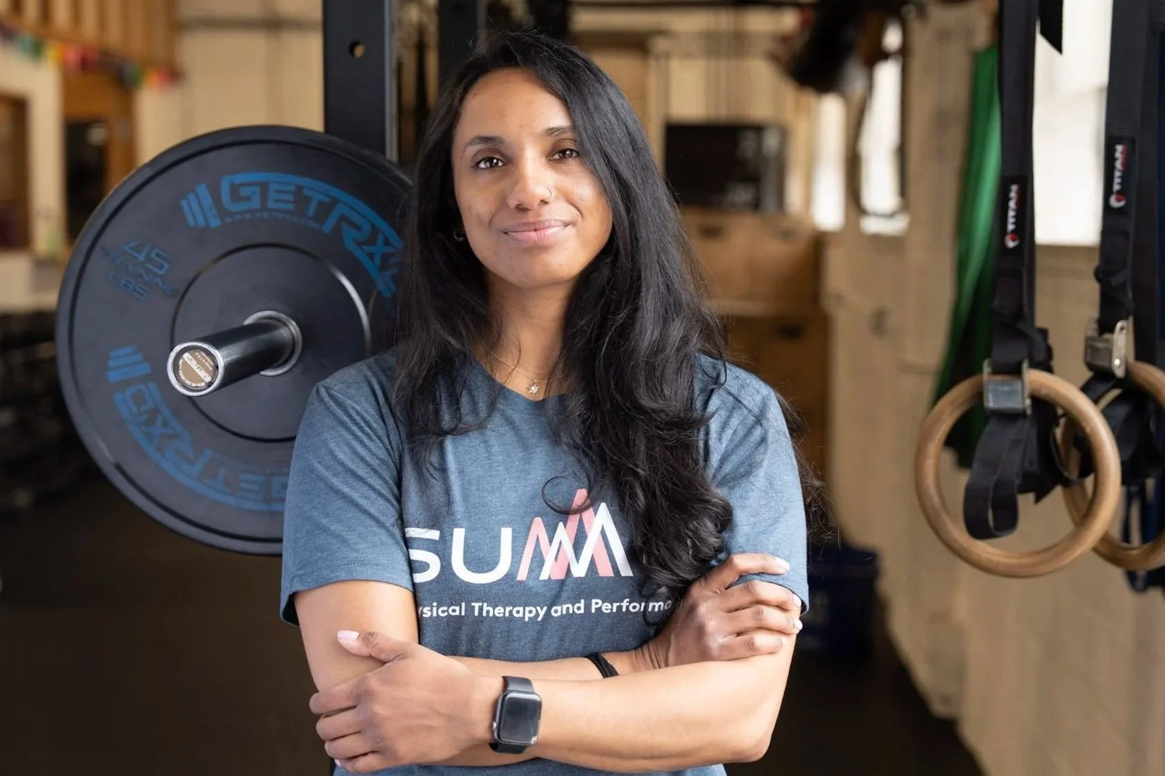 A woman with long dark hair smiling with arms crossed standing in a gym near gymnastic rings.