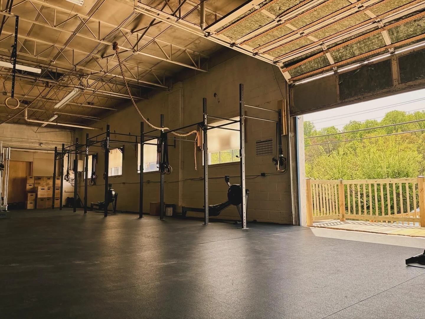 Empty gym with fitness equipment, open garage door showing outdoor wooden deck and green trees.
