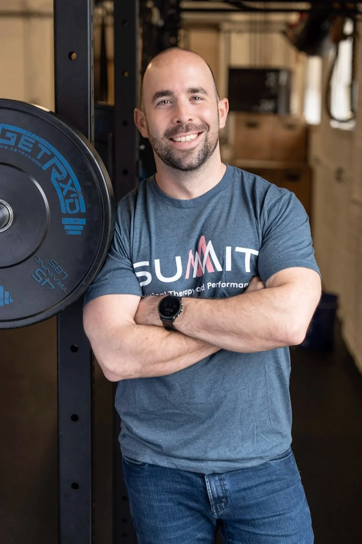 A man with a beard and a shaved head smiling with arms crossed, wearing a blue T-shirt with the word 'SUMMIT' printed on it, standing in a dimly lit gym or fitness center.