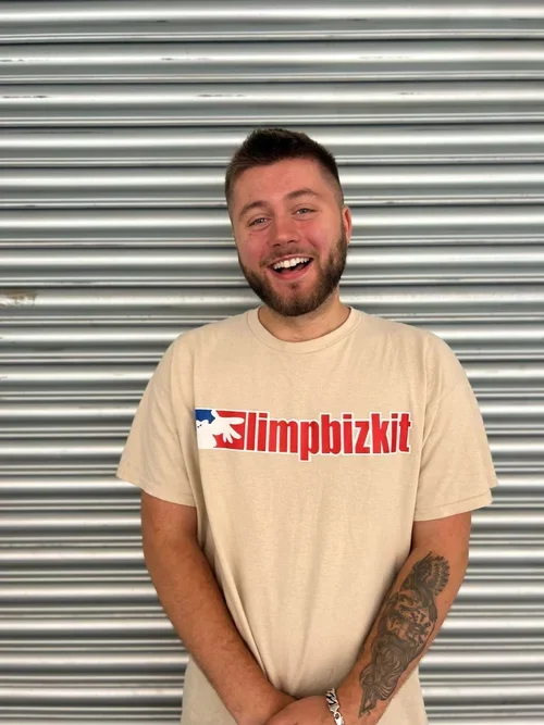 A young man with a beard and short hair, smiling, standing in front of a metal shutter. He is wearing a beige t-shirt with the text 'limpbizkit' in red and blue, featuring a graphic of a hand holding a microphone.