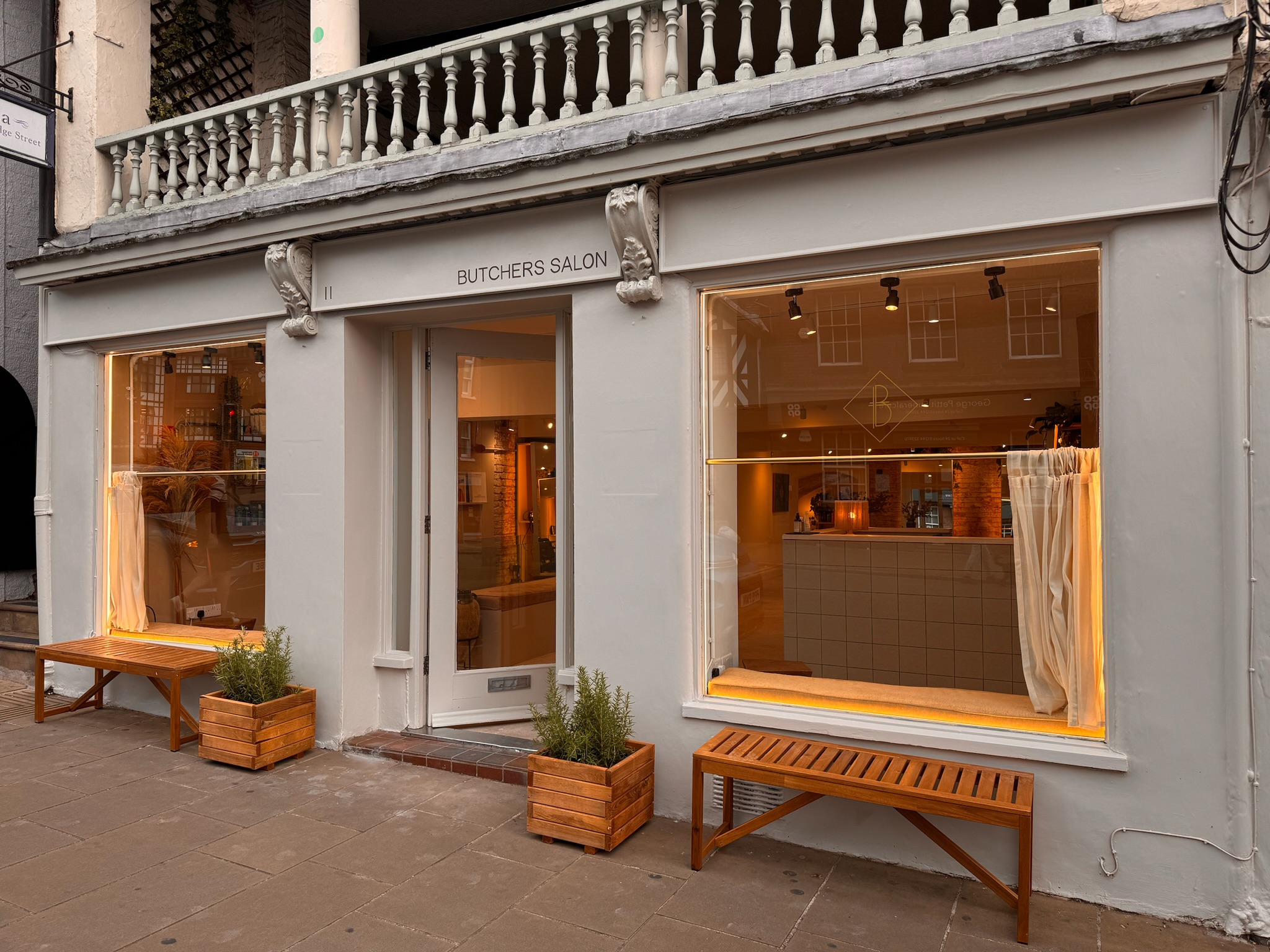 Exterior of a white storefront with large windows, labeled 'Butchers Salon,' featuring warm lighting inside, orange curtains, wooden benches, and potted plants outside.