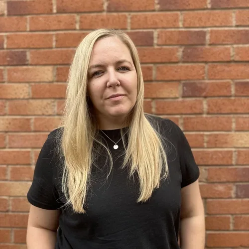 A woman with blonde hair wearing a black t-shirt standing in front of a brick wall.