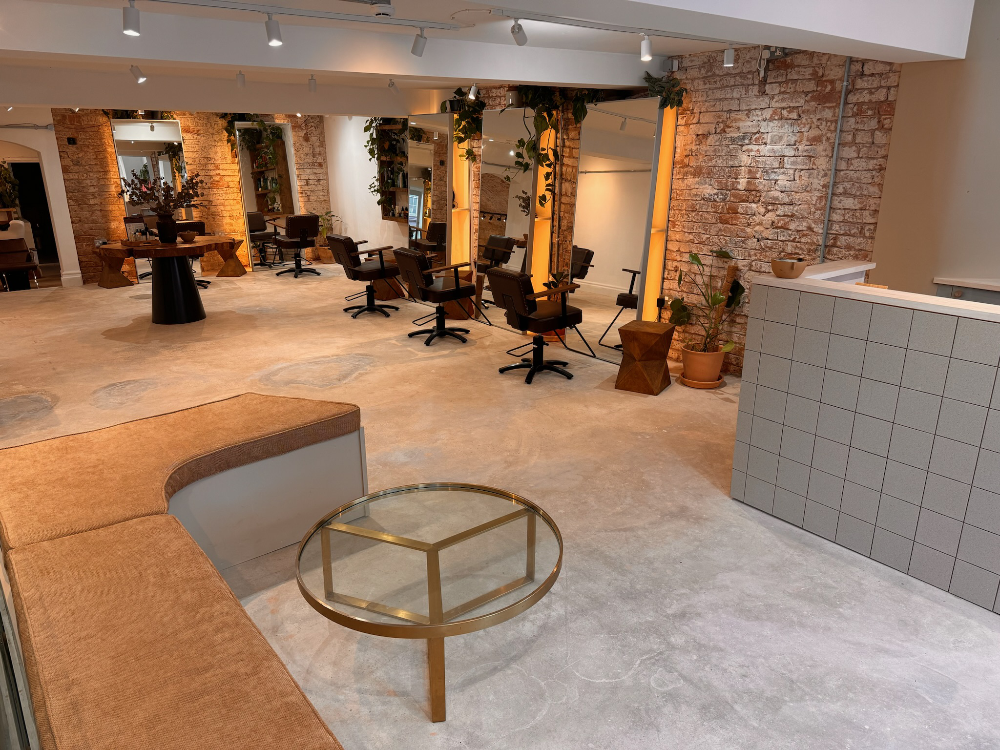 Empty modern salon with hair styling chairs in front of mirrors, exposed brick walls, a seating area with a brown sofa and glass coffee table, and potted plants.