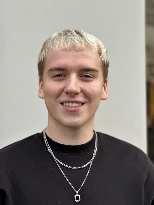 A young man with blonde hair smiling, wearing a black shirt and layered silver necklaces, standing against a plain light background.