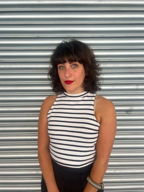 A woman with shoulder-length dark hair and bangs, wearing a sleeveless, high-neck, striped top and black pants, standing in front of a metallic, horizontal roller shutter.