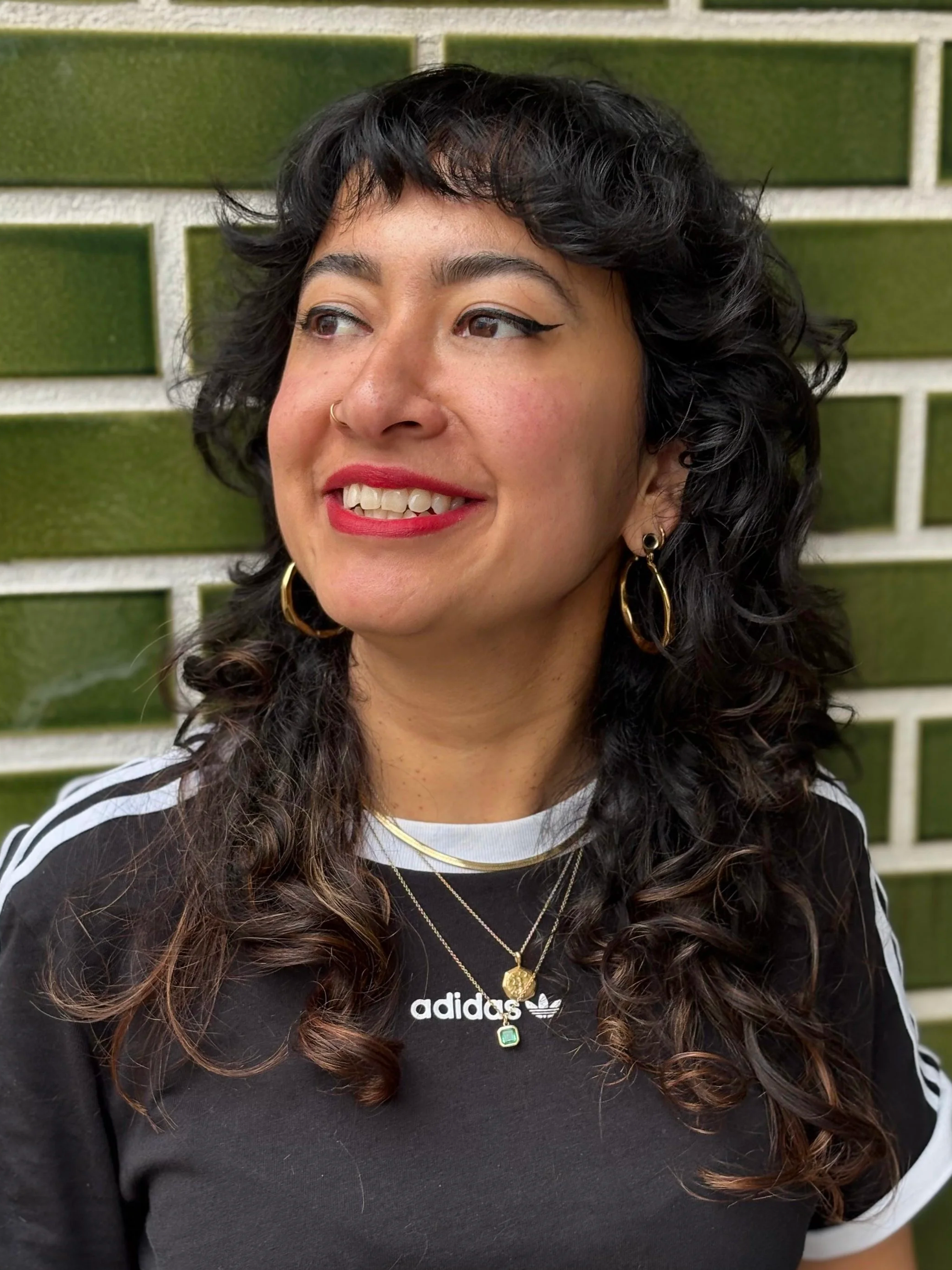 A woman with black curly hair, wearing a black Adidas shirt with white stripes, gold hoop earrings, layered necklaces, and red lipstick, standing in front of a green and white brick wall.