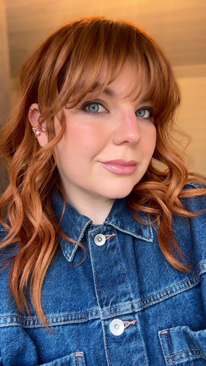Close-up of a woman with red hair, blue eyes, and fair skin, wearing a denim jacket and a sparkly earring. She has a neutral expression and is looking slightly to the side.