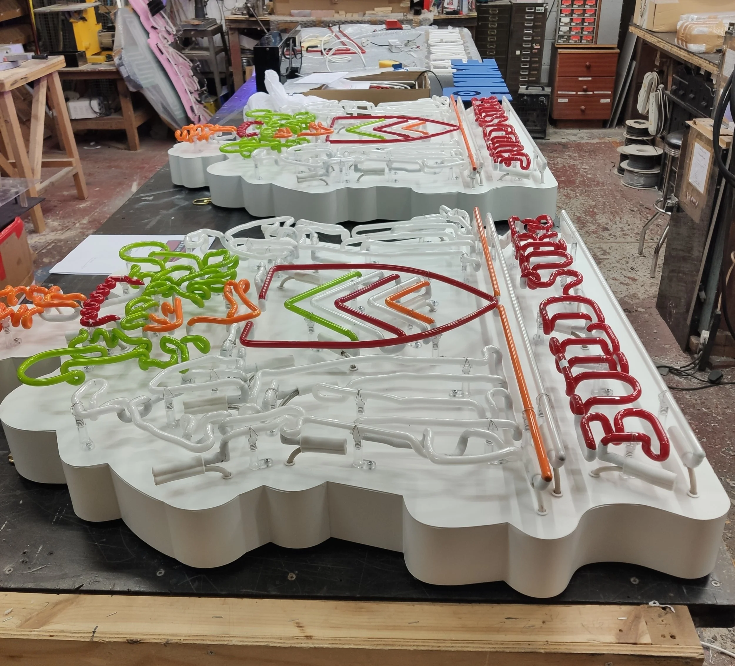 Colorful neon signs in process of being assembled on a workshop table, with various tools and drawers in the background.