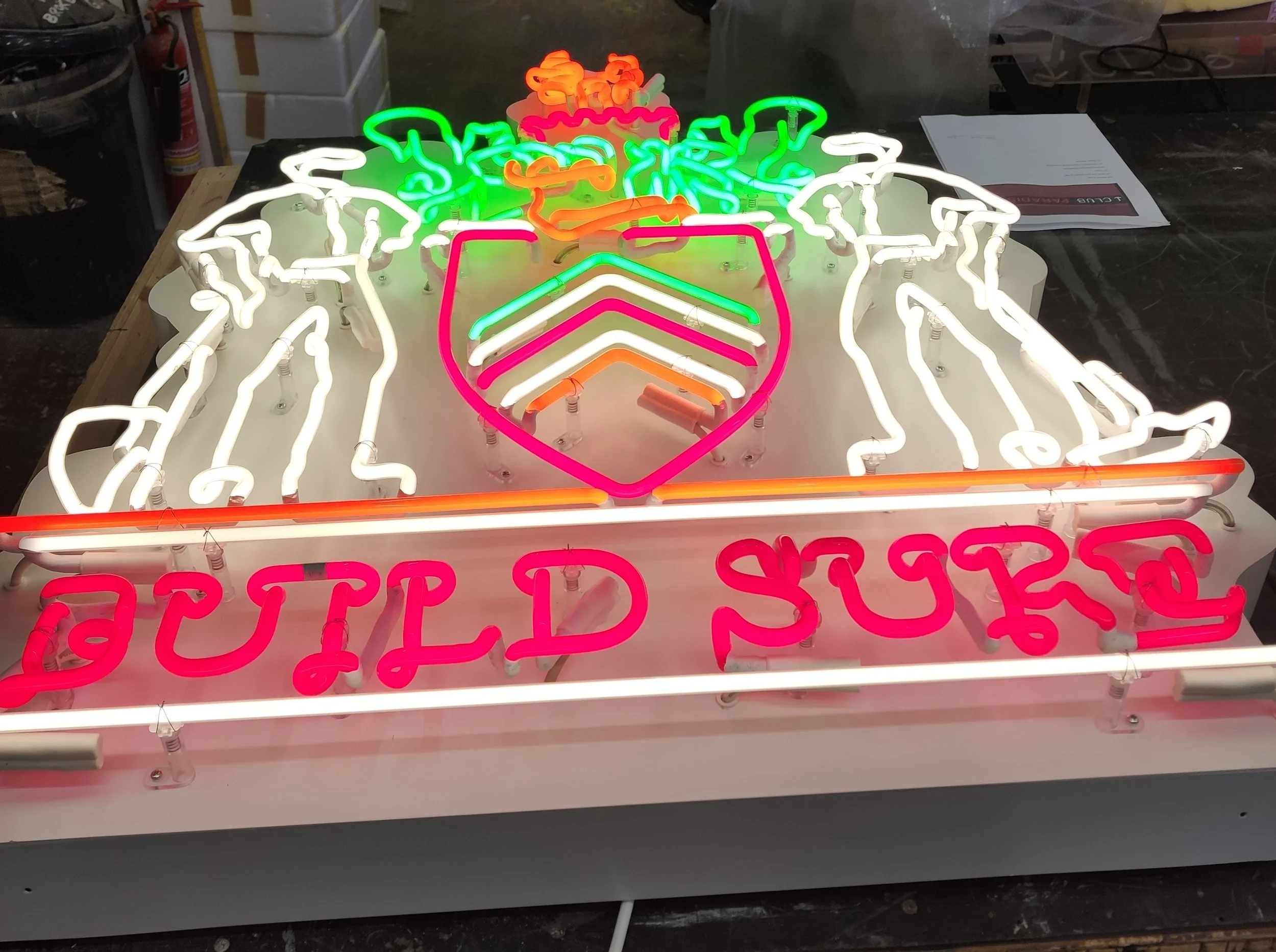 Colorful neon sign with a shield, a crown, and the words 'BUILD YOUR' in pink, with various decorative elements.