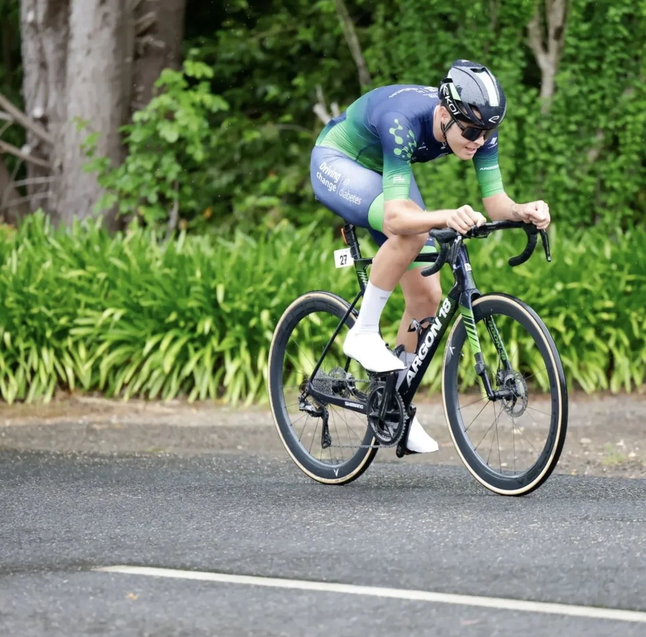 A cyclist wearing a blue and green uniform riding a black Argon 18 road bike on a paved road with green bushes and trees in the background.