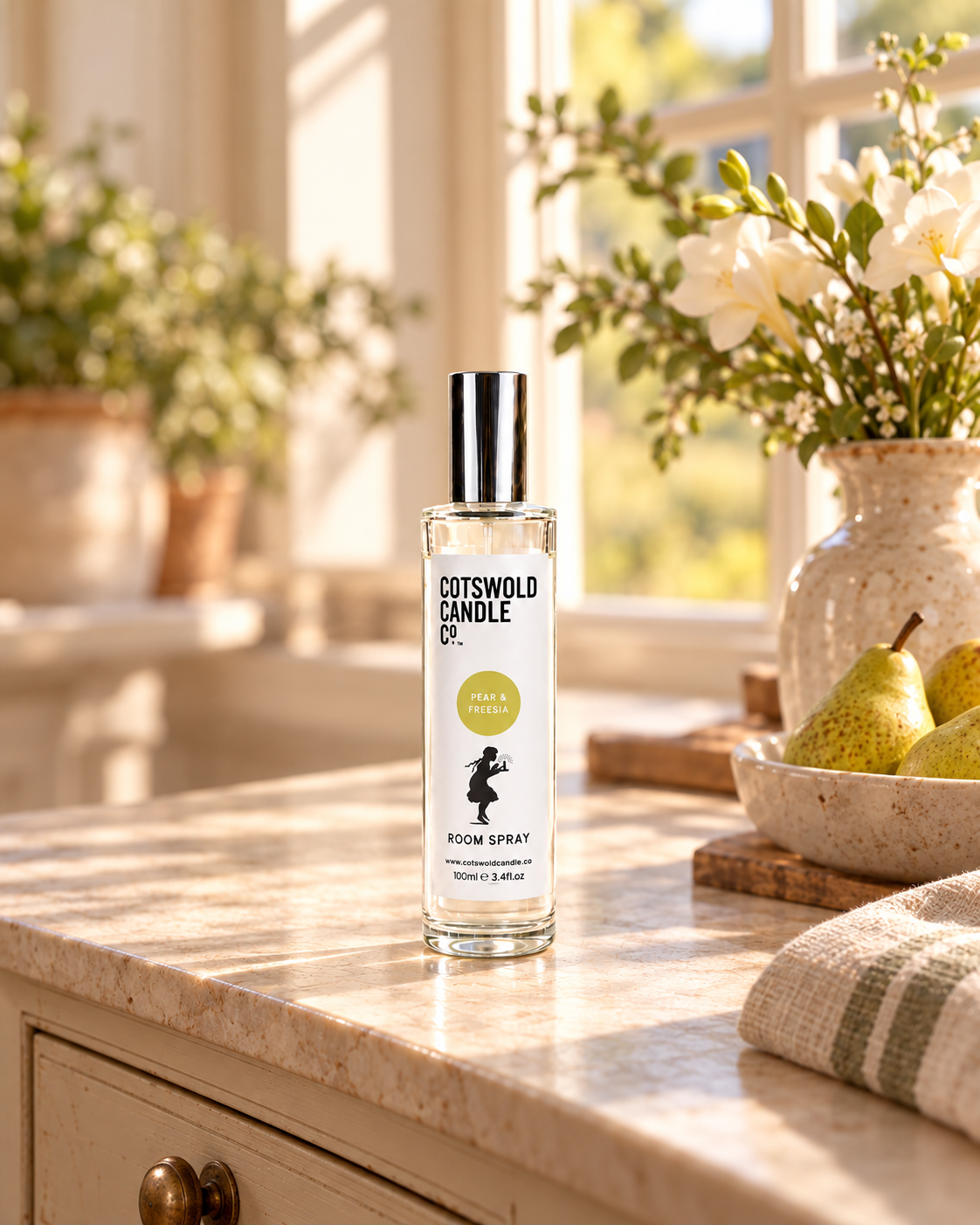 Pear & Freesia room spray by Cotswold Candle Co., clear glass 100ml bottle with silver cap, soft yellow green fragrance circle and girl logo, in a softly sunlit home setting.