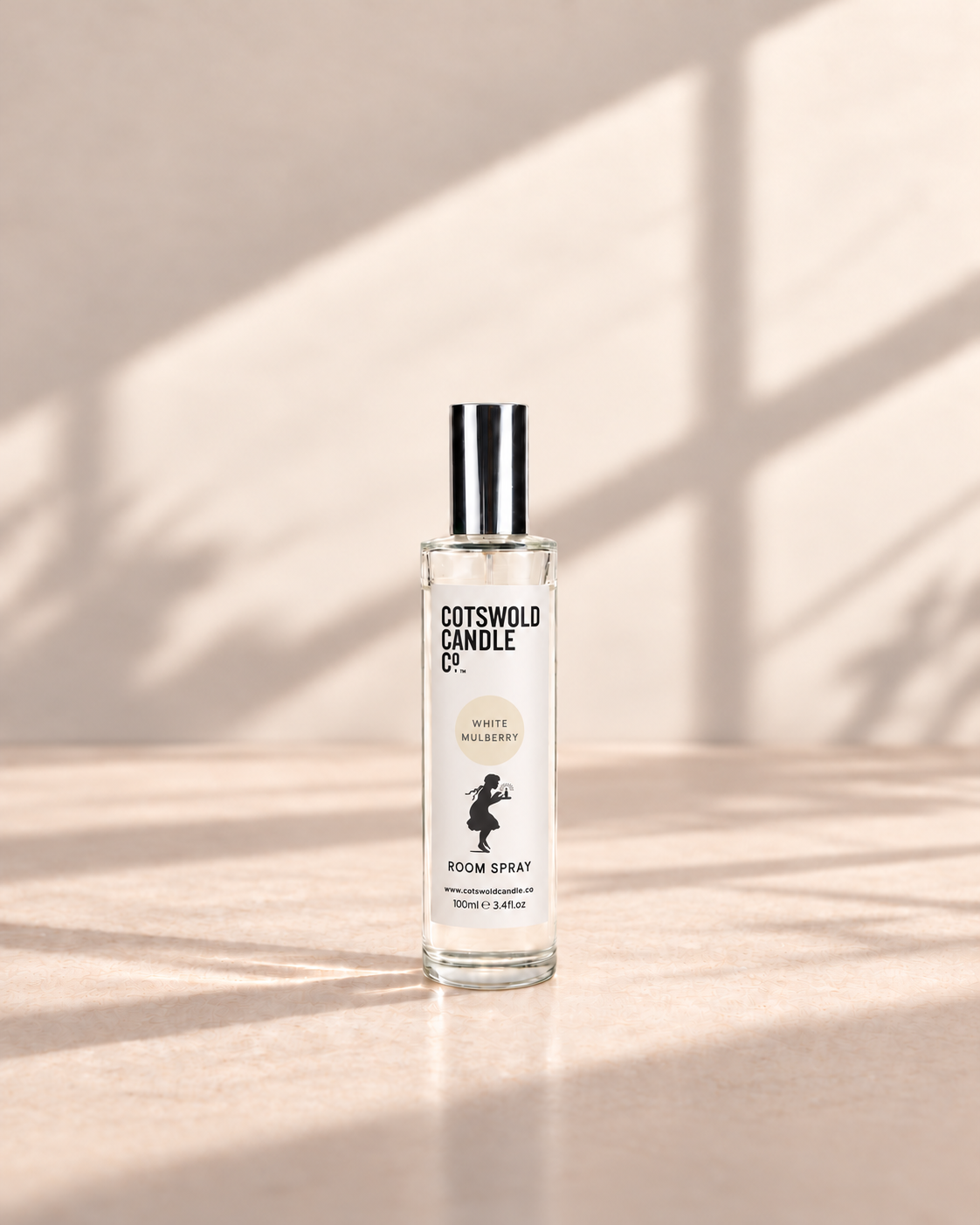 White Mulberry room spray by Cotswold Candle Co., clear glass 100ml bottle with silver cap, cream fragrance circle and girl logo, on a warm neutral studio background.