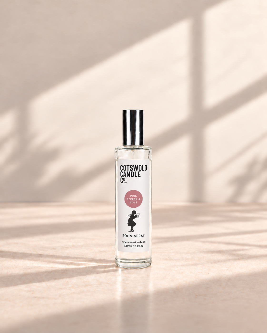 Pink Pepper & Rose room spray by Cotswold Candle Co., clear glass 100ml bottle with silver cap, dusty pink fragrance circle and girl logo, on a warm neutral studio background.