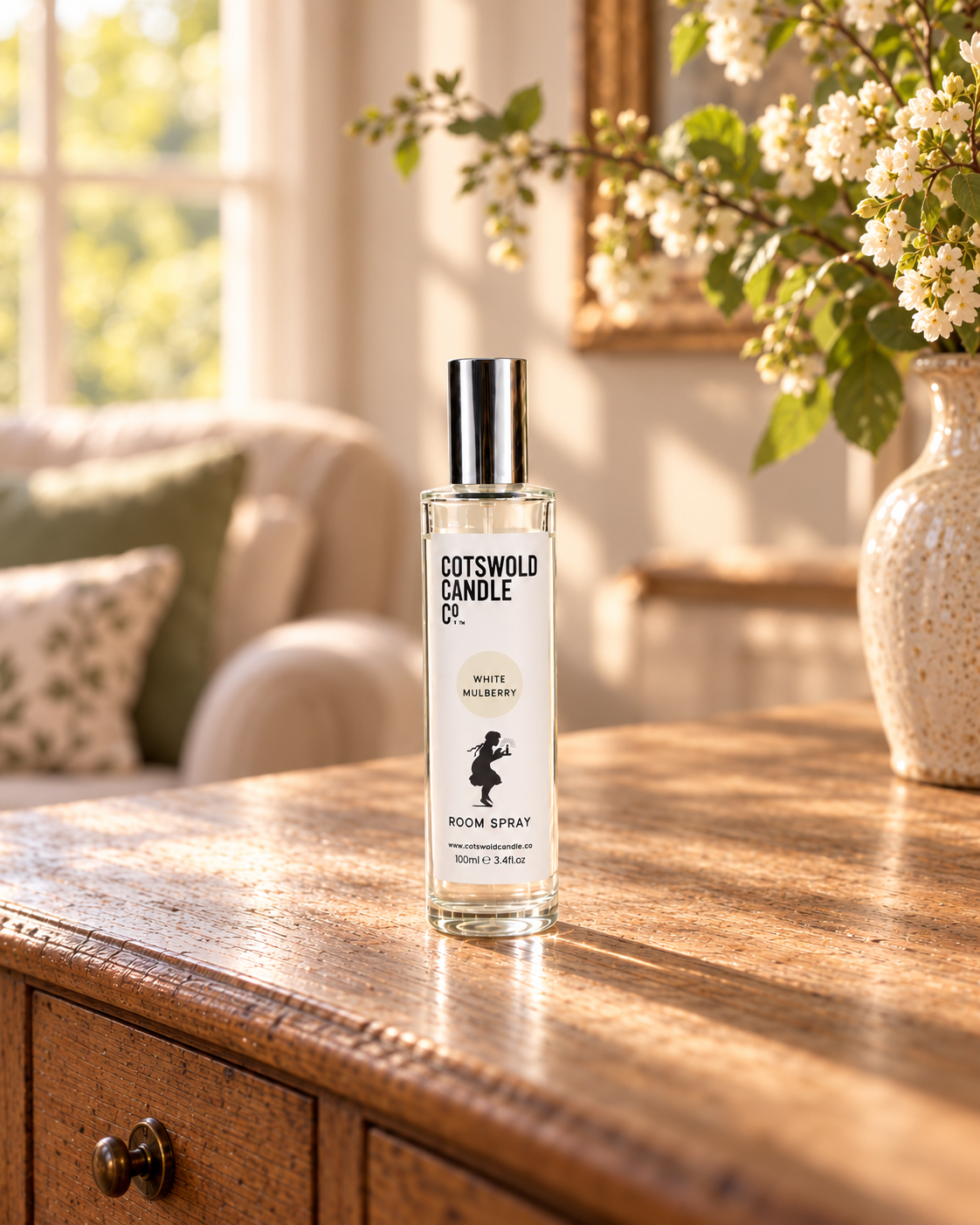 White Mulberry room spray by Cotswold Candle Co., clear glass 100ml bottle with silver cap, cream fragrance circle and girl logo, in a softly sunlit home setting.